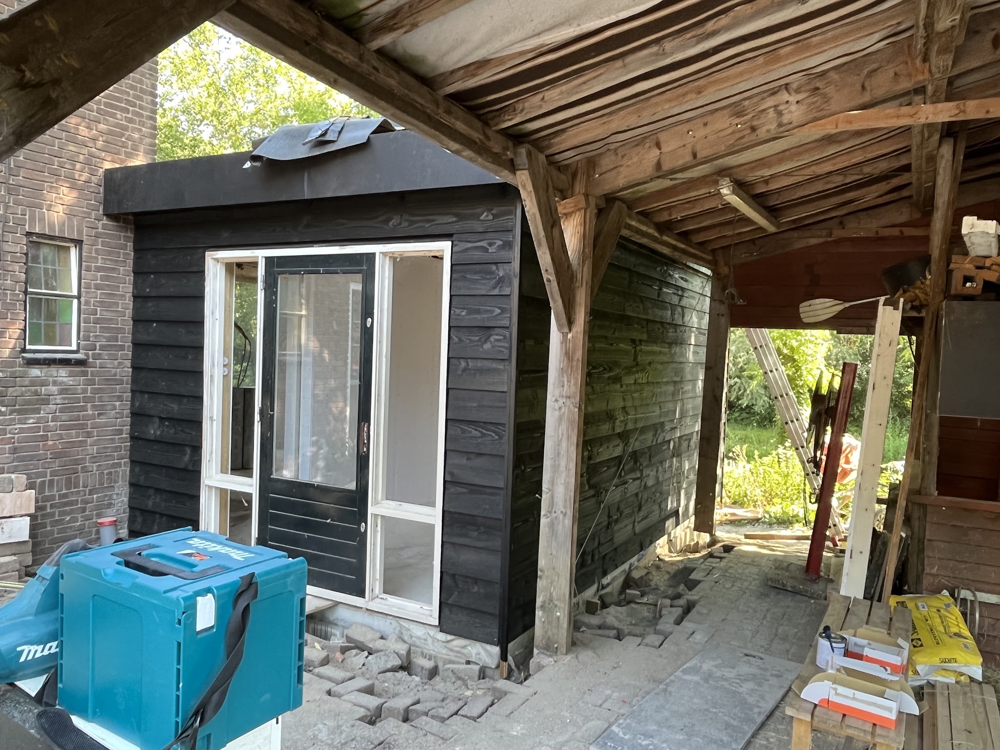 Zwarte houten tuinkamer in aanbouw onder een oude overkapping, met gereedschapskoffer op de voorgrond